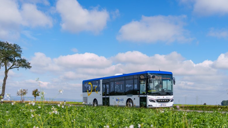 Unterwegs mit den LEOpoldi-Linienbussen, © NÖVOG/Bollwein Blaue-weißer Linienbus auf einer Straße vor grünem Feld unter blauem Himmel mit weißen Wolken