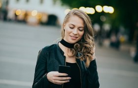 Junge Frau mit lockigem Haar und schwarzer Jacke liest lächelnd eine Nachricht auf ihrem Smartphone