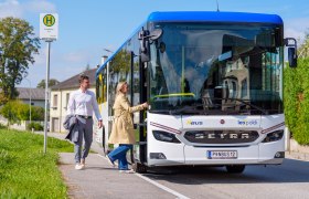Weißer Setra-Bus mit blauem Dach an Haltestelle, zwei Personen steigen ein, sonniger Tag, ländliche Umgebung.