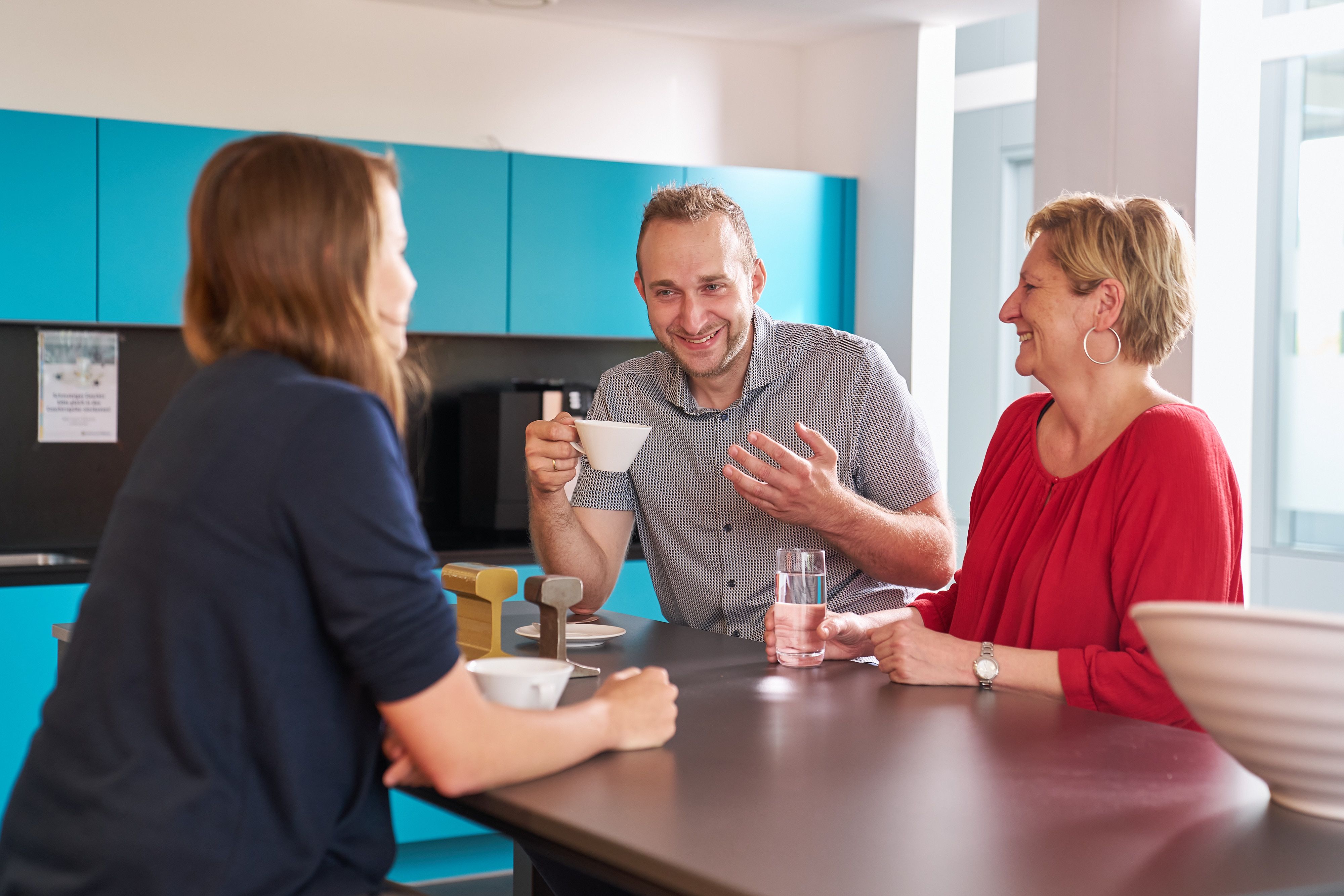 Drei Mitarbeitende unterhalten sich bei Kaffee und Wasser in einer Büroküche.