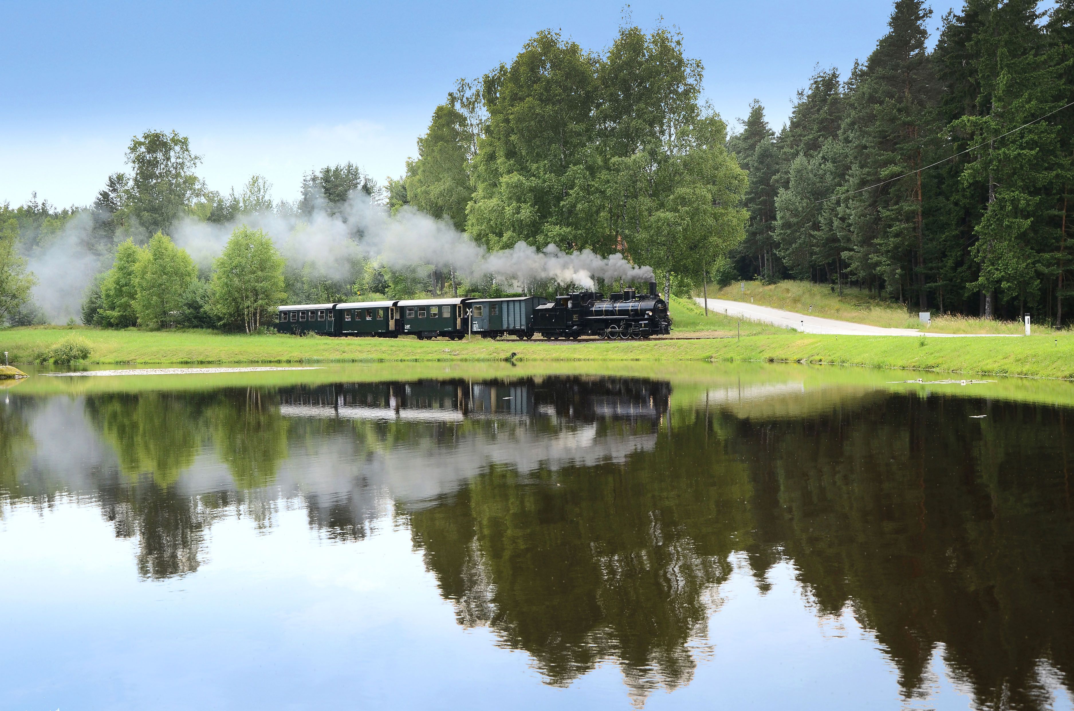 Dampfzug Waldviertelbahn im Sommer. Im Vordergrund sieht man einen Teich, im Hintergrund die typische und urtümliche Landschaft des Waldviertels mit seinen bewaldeten Hügeln.