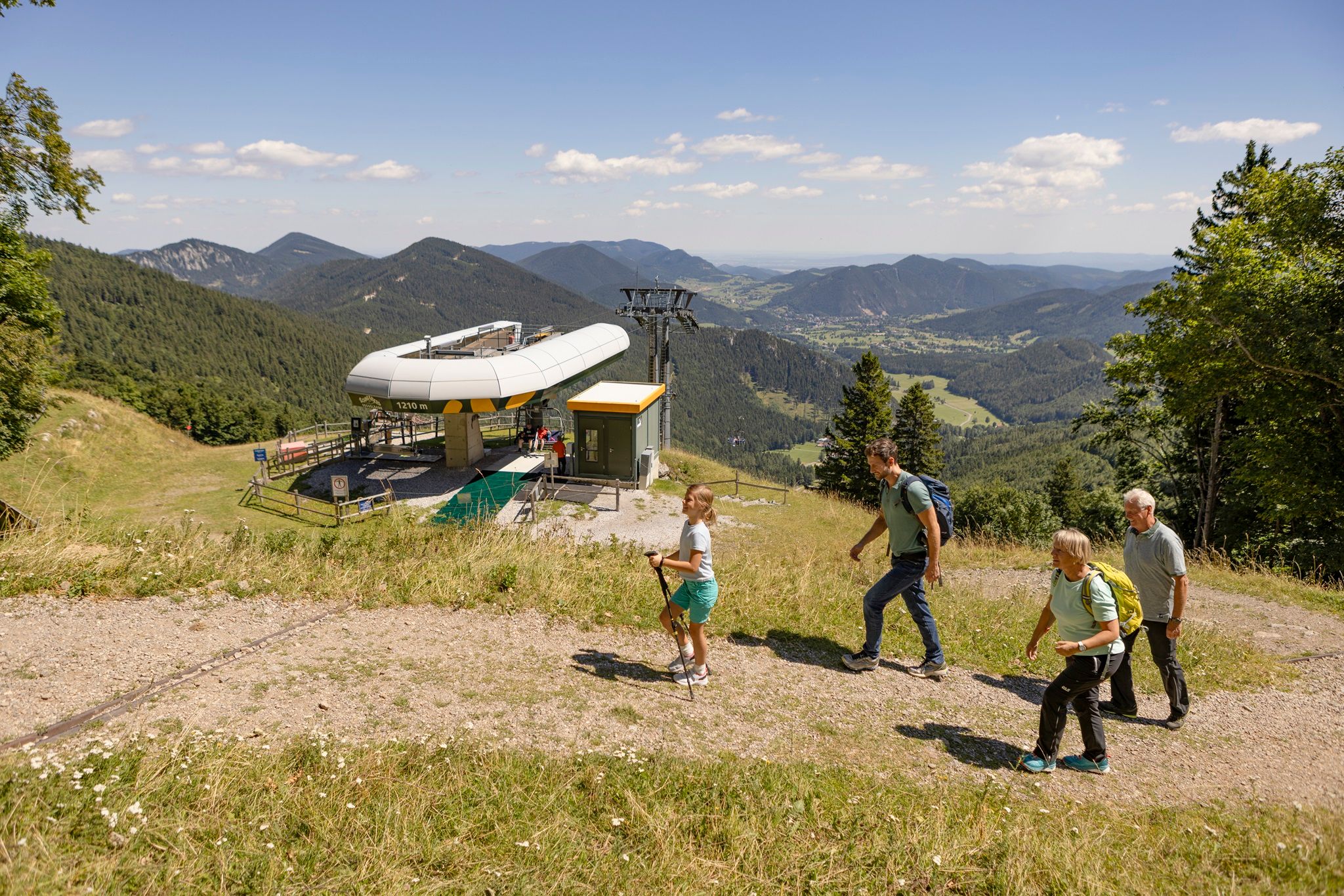 Bergstation der Schneeberg Sesselbahn. Eine Familie wandert gemütlich den Berg hinauf, im Hintergrund sieht man die Bergstation der Sesselbahn und das alpine Panorama der umliegenden Bergwelt.