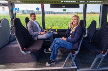 Zwei Personen sitzen sich in einem Bus gegenüber, eine mit Laptop auf dem Schoß, die andere telefoniert, beide tragen Blazer und Jeans.
