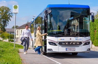 Weißer Setra-Bus mit blauem Dach an Haltestelle, zwei Personen steigen ein, sonniger Tag, ländliche Umgebung.