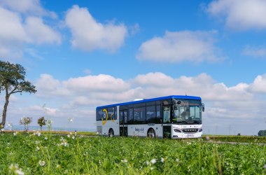 Blaue-weißer Linienbus auf einer Straße vor grünem Feld unter blauem Himmel mit weißen Wolken