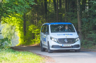 Weißer Mercedes-Van mit blauem Schriftzug 'leopoldi' fährt auf schmaler Straße durch bewaldete Landschaft.