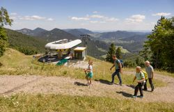 Bergstation der Schneeberg Sesselbahn. Eine Familie wandert gemütlich den Berg hinauf, im Hintergrund sieht man die Bergstation der Sesselbahn und das alpine Panorama der umliegenden Bergwelt.