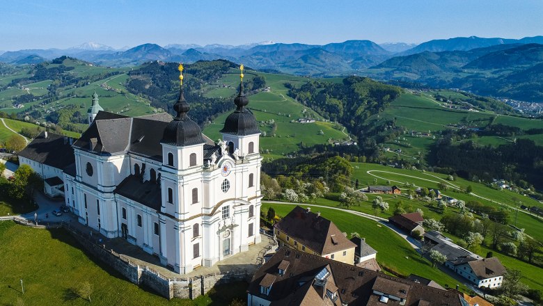 Barocke Kirche mit zwei Türmen auf einem Hügel, umgeben von Häusern und grünen Wiesen, im Hintergrund Berge unter blauem Himmel.
