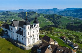Barocke Kirche mit zwei Türmen auf einem Hügel, umgeben von Häusern und grünen Wiesen, im Hintergrund Berge unter blauem Himmel.