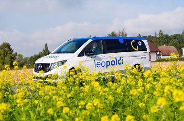 Weißer Mercedes-Van mit blauer und gelber Schrift 'leopoldi' auf der Seite, steht auf einem Feld mit gelben Blumen, im Hintergrund Bäume und ein Haus.