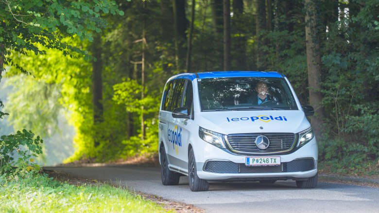 Weißer Mercedes-Van mit blauem Schriftzug 'leopoldi' fährt auf schmaler Straße durch bewaldete Landschaft.