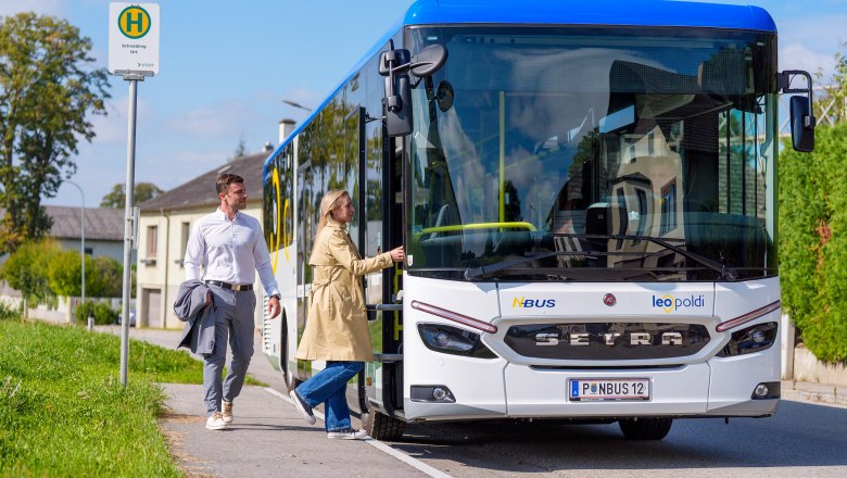 Weißer Setra-Bus mit blauem Dach an Haltestelle, zwei Personen steigen ein, sonniger Tag, ländliche Umgebung.