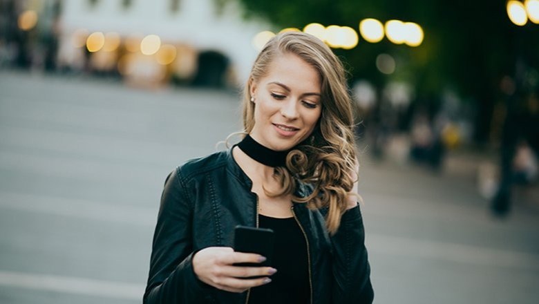 Junge Frau mit lockigem Haar und schwarzer Jacke liest lächelnd eine Nachricht auf ihrem Smartphone